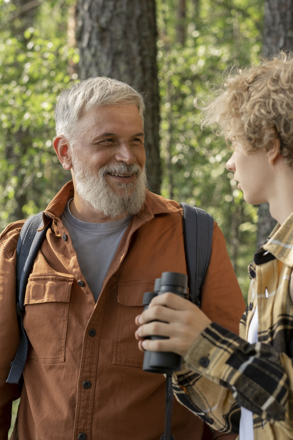 A smiling older man in an orange jacket with a gray beard talks to a younger person holding binoculars, surrounded by a lush forest setting in apache junction az