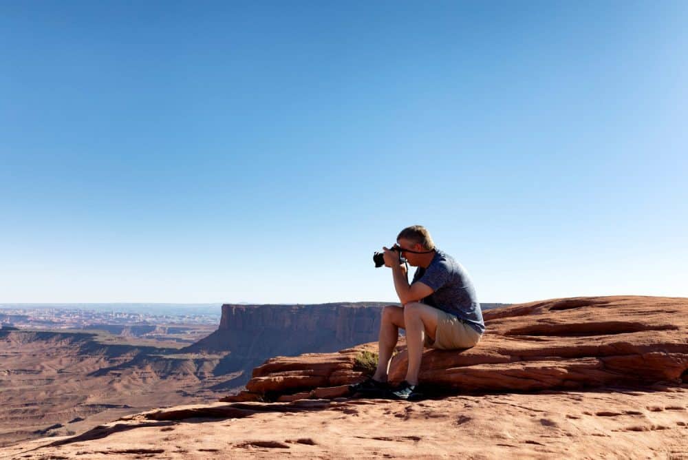 A man sitting on red rocks with a camera, photographing a vast canyon landscape under a clear blue sky in apache junction az