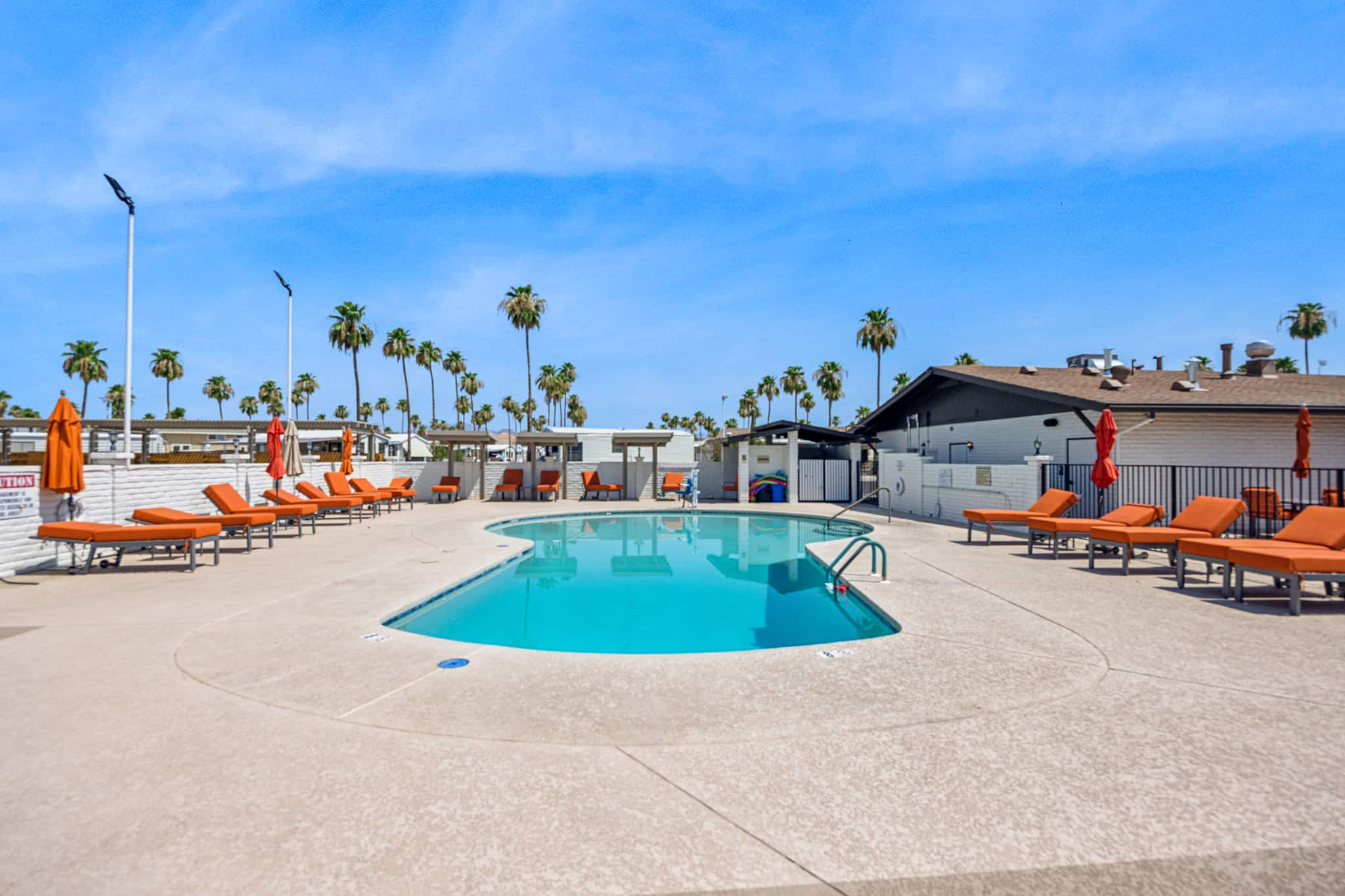 A kidney-shaped pool with orange lounge chairs and umbrellas, set against a backdrop of palm trees and a bright blue sky in apache junction az