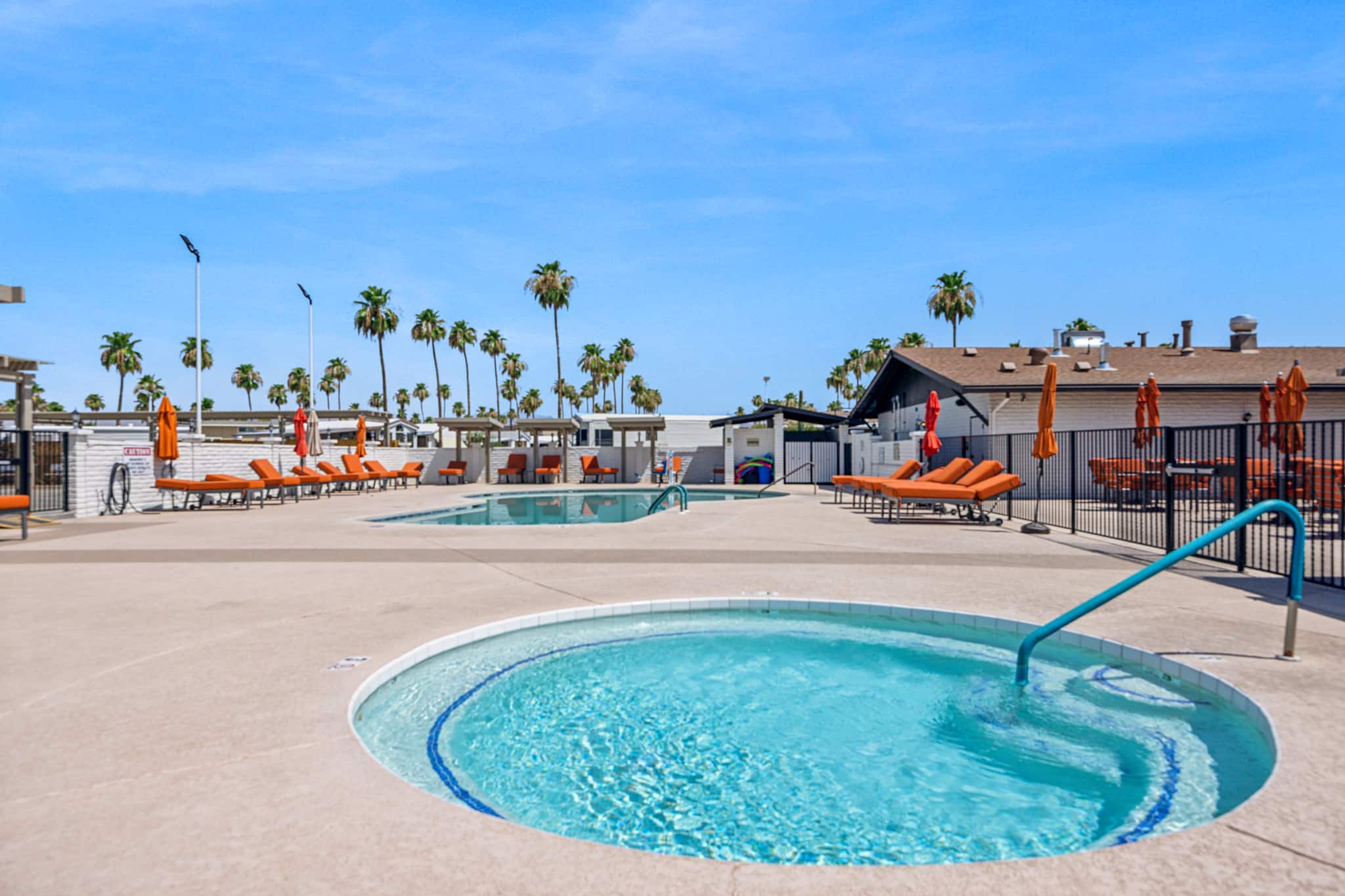 A pool and hot tub area with orange lounge chairs and umbrellas, surrounded by palm trees and a sunny blue sky in apache junction az
