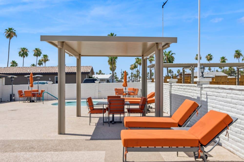 A poolside cabana with orange lounge chairs and tables, surrounded by white fencing and palm trees under a bright blue sky in apache junction az