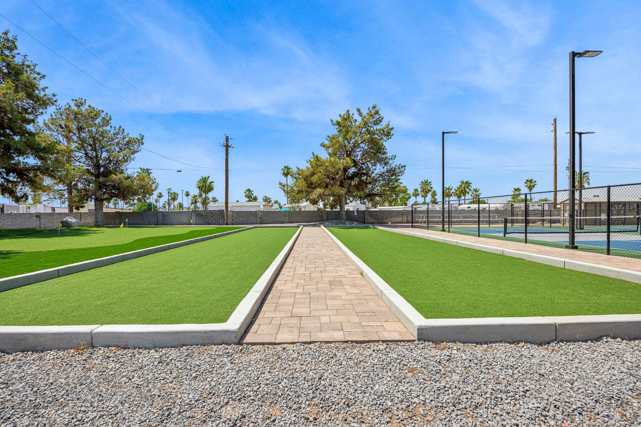 A bocce ball court with two lanes of artificial turf, surrounded by a landscaped area and a paved path, with a blue sky overhead in apache junction az