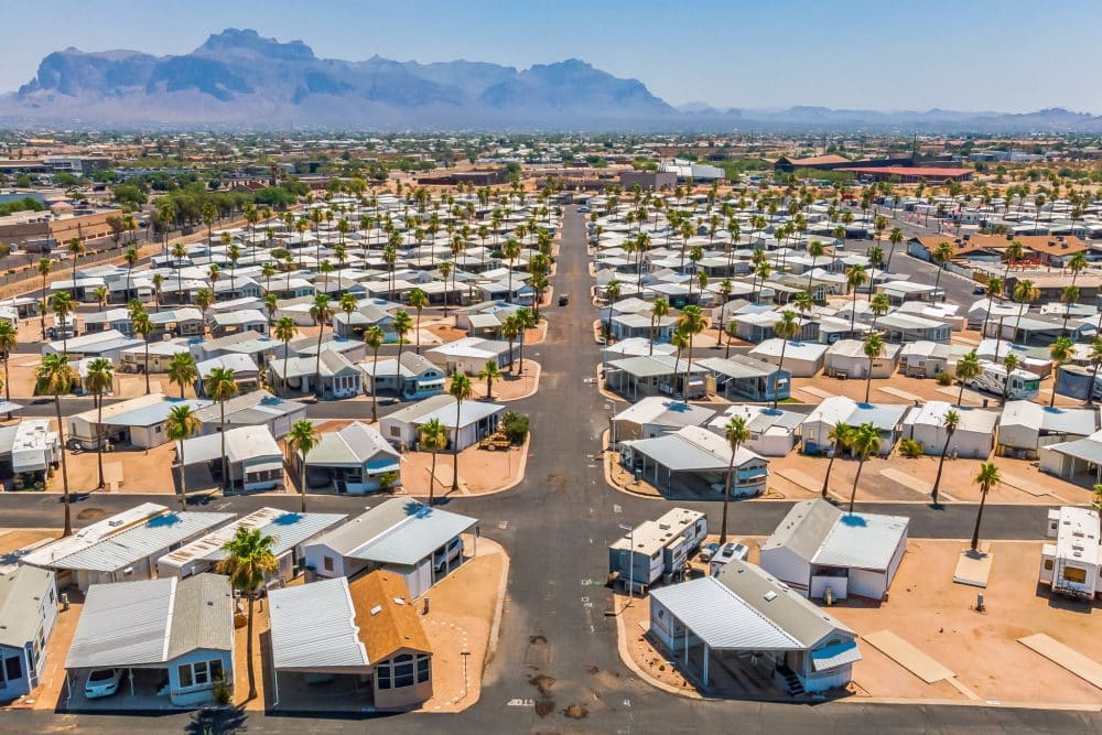 An aerial view of a large residential community with rows of homes, palm trees, and mountains in the distance under a sunny sky in apache junction az